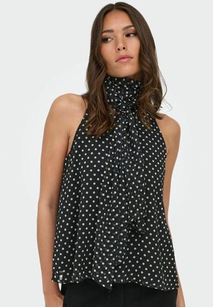Femme aux cheveux bruns ondulés portant un haut noir sans manches à pois blancs avec un détail de noeud au col montant, regardant sur le côté.