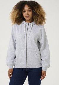 Pelēks zip-up kapucis, izgatavots no mīkstas auduma. Iespējas ietver oderētu kapuci, regulējamus auklas un priekšējos kabatas. Pieguļošas piedurknes un brīva silueta.