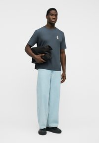 Bărbat purtând un tricou gri închis cu un mic grafic și blugi evazați bleu deschis, ținând o geantă neagră pliată.