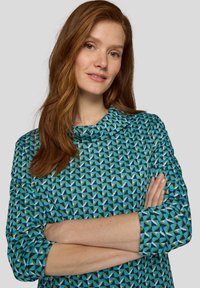 Frau mit langen roten Haaren, die ein blaues Langarmoberteil mit geometrischem Muster trägt, steht mit verschränkten Armen vor einem einfarbigen grauen Hintergrund.