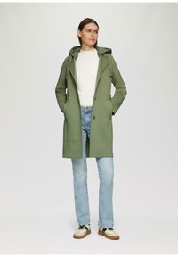 Grüne Kapuzenjacke aus weichem Stoff, mit lockerer Passform, Fronttaschen und einem einzelnen Knopfverschluss. Über einem weißen Pullover und Jeans getragen.
