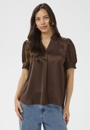 Femme aux longs cheveux ondulés portant des boucles d'oreilles créoles en or, un chemisier marron en satin à manches courtes et un jean bleu clair, debout devant un fond uni.