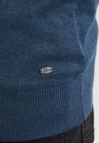 Pulover albastru cu textură de tricot cu nervuri. Prezintă un mic patch cu logo argintiu etichetat "Petrol" în partea stângă jos.