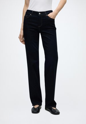 Personne portant un jean noir droit avec la main dans la poche, un haut blanc sans manches et des ballerines noires sur fond blanc uni.