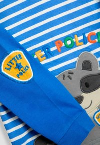 Kék csíkos ing, hímzett mosómedve karakterrel, színes betűkkel írt "SZEMÉTÜGYI EGYSÉG" felirattal, és egy kék jelvénnyel az ujjon.