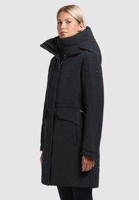Cămașă lungă, izolată, de culoare neagră, cu un guler înalt, fermoar frontal și buzunare largi. Design elegant cu branding subtil și țesătură texturată.