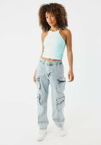 Top corto a costine blu chiaro e jeans cargo a lavaggio chiaro con molte tasche con zip. Modello che indossa scarpe da ginnastica bianche, in piedi su uno sfondo bianco.