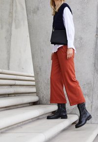 Bottes de combat noires, pantalon ample couleur rouille, et un gilet tricoté bleu marine porté sur une chemise blanche, sur fond de béton avec des marches courbes.