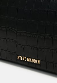 Svart krokodillepreget håndveske med strukturert overflate. Har en gull "Steve Madden"-logo nederst. Elegant, rektangulær design.