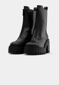 Paire de bottes à plateforme en cuir noir avec talons épais et fermetures éclair devant sur un fond clair uni.