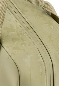 Vattenresistent beige väska med texturerad yta, synliga vattendroppar och kontrasterande tan-sömmar och remmar, med en strömlinjeformad design.