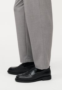 Schwarze Leder-Slipper getragen zu grauen, maßgeschneiderten Hosen vor einem schlichten weißen Hintergrund.