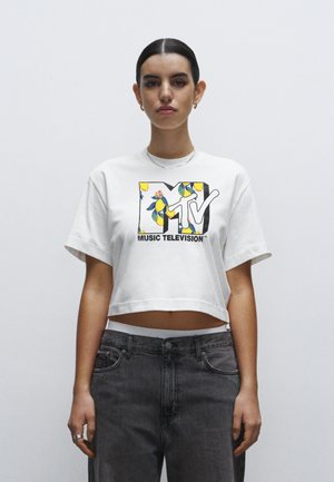Cropped white t-shirt with a colorful MTV graphic featuring floral and lemon patterns. Model pairs it with faded gray jeans.