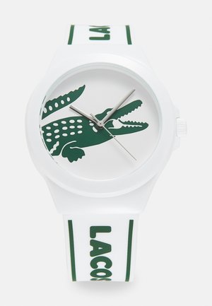 Baltas gumijas pulkstenis ar zaļu krokodila grafiku uz ciparnīcas un zaļu "LACOSTE" tekstu uz siksniņas. Iespējas: sudrabaini rādītāji un gluda apdare.