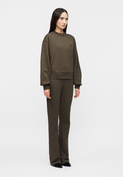 Γυναίκα με μακριά σκουρόχρωμα μαλλιά, φοράει χαλαρό καφέ πουλόβερ και αντίστοιχο παντελόνι, στέκεται μπροστά σε απλό φωτεινό φόντο.