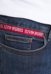 Vaqueros de denim con un lavado azul y costuras naranjas. Una etiqueta de tela roja dice "ELEVEN DEGREES" en la cinturilla. Detalle del bolsillo delantero visible.