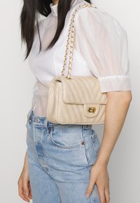 Beige gesteppte Lederhandtasche mit einem goldenen Kettenriemen und Verschluss, rechteckige Form, präsentiert vor einem hellen Hintergrund mit einem Modell.