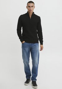 Pull à col zippé noir en tricot avec un motif texturé, associé à un jean droit bleu et des baskets basses noires.