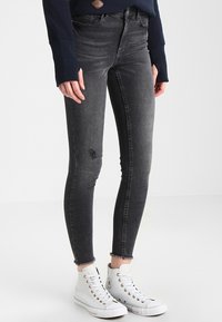 Jeans skinny negros con detalle desgastado en las rodillas y dobladillo deshilachado. El material parece denim con una textura suave. Llevados con zapatillas altas blancas.