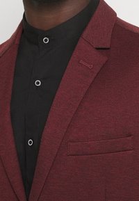 Bordowa kurtka z teksturowanej tkaniny, o ostrych klapach i kieszeni na piersi; zestawiona z czarną koszulą na guziki z okrągłymi guzikami.