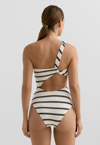 Egyrészes krémszínű fürdőruha tengerészkék vízszintes csíkokkal, egyvállpánttal és nyitott hátkivágással.