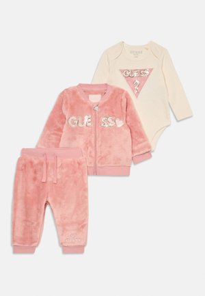 Tryse dalykai kūdikiams: minkštas rožinis užtrauktuku užsegamas švarkas su "GUESS" logotipu, suderintos rožinės kelnės ir kreminės spalvos kūdikio bodysuitas su rožinės spalvos trikampio raštu.
