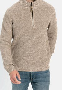 Beige gestrickter Pullover mit einem halben Reißverschluss, strukturiertem Stoff und gerippten Bündchen. Über blauen Denim-Jeans getragen, was ein lässiges Design zeigt.