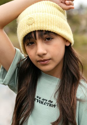 Geel gebreide muts met ribbelstructuur en logo patch. Lichtgroen T-shirt met zwarte geborduurde tekst. Lang, donker haar.