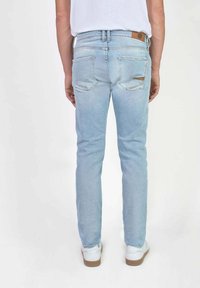 Vaqueros de mezclilla azul claro con corte ajustado, bolsillos traseros, ligero desgaste y una etiqueta de cuero marrón en la cintura. Zapatillas blancas visibles.