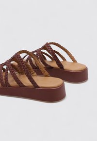 Sandalias trenzadas marrones con un diseño de banda ancha, parte superior texturizada y suela platillo. El forro interior suave contrasta con la suela exterior.