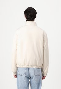 Beige, letvægtjakke med høj krave, elastiske manchetter og kant, der har en glat tekstur og minimalistisk design.