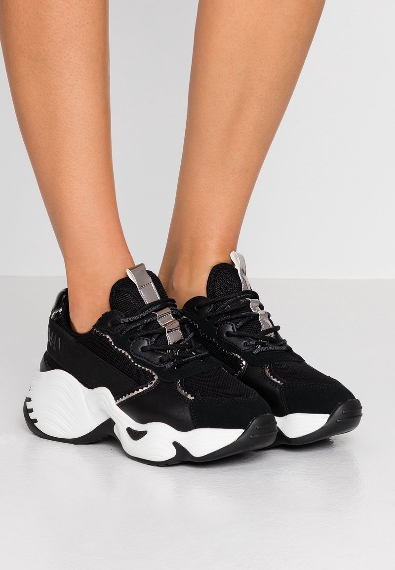 Black sneakers with mesh and suede material, featuring white accents and a thick white sole. Silver hardware on top lace area.