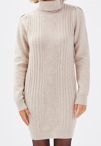 Vestito a collo alto in maglia cable beige con maniche allungate e polsini a coste. Il motivo testurizzato si sviluppa verticalmente lungo il centro.
