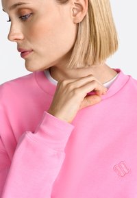 Sweatshirt rose avec des poignets et un col côtelés. Présente un petit logo en relief. Vue rapprochée de la texture du tissu et de la position de la main.