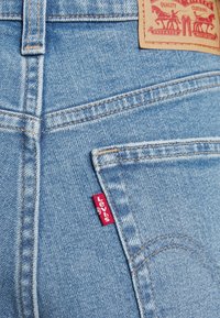 Poche arrière d’un jean denim bleu clair avec étiquette rouge de la marque Levi’s et pièce en cuir beige avec logo et détails de couture.