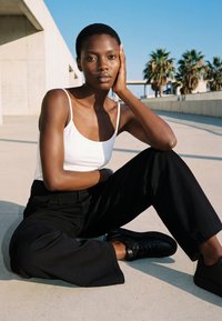 Jeune personne aux cheveux courts, vêtue d’un débardeur blanc et d’un pantalon noir, assise sur le sol en béton à l’extérieur, reposant la tête sur la main, palmiers en arrière-plan.