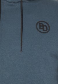 Sudadera con capucha en tejido azul oscuro, con un logo negro, cordones ajustables negros y un acabado texturizado.