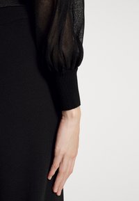 Bras en robe noire avec manche ballon transparente et poignet côtelé, montrant une main pâle reposant sur un fond en tissu noir.