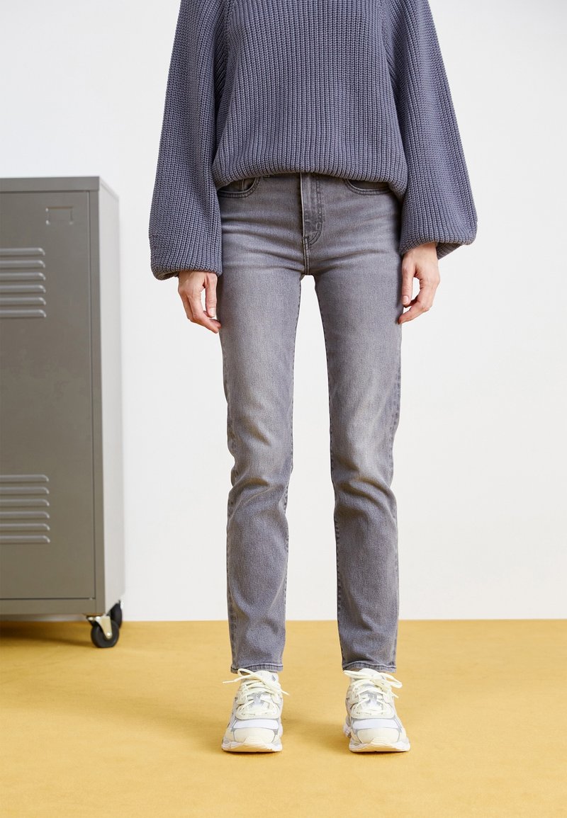 Personne debout portant un jean gris, des baskets blanches et un pull en tricot violet, à côté d'un casier métallique gris sur un sol jaune.