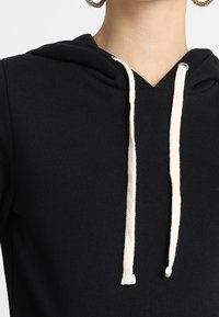 Sudadera con capucha negra y cordones blancos, llevada por una persona que muestra el pecho superior y el cuello, con grandes pendientes de aro visibles.
