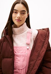 Chaqueta de plumas marrón, peto de correa de pana color rosa claro con un bolsillo y detalles en plata, y un suéter de punto de cuello alto con motas multicolores.