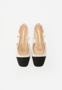 Beige slinga kurpes ar melnu tekstūrētu deguna daļu, gludu iekšējo zoli, regulējamu siksniņu un noapaļotu formu. Vienkāršs, elegants dizains.