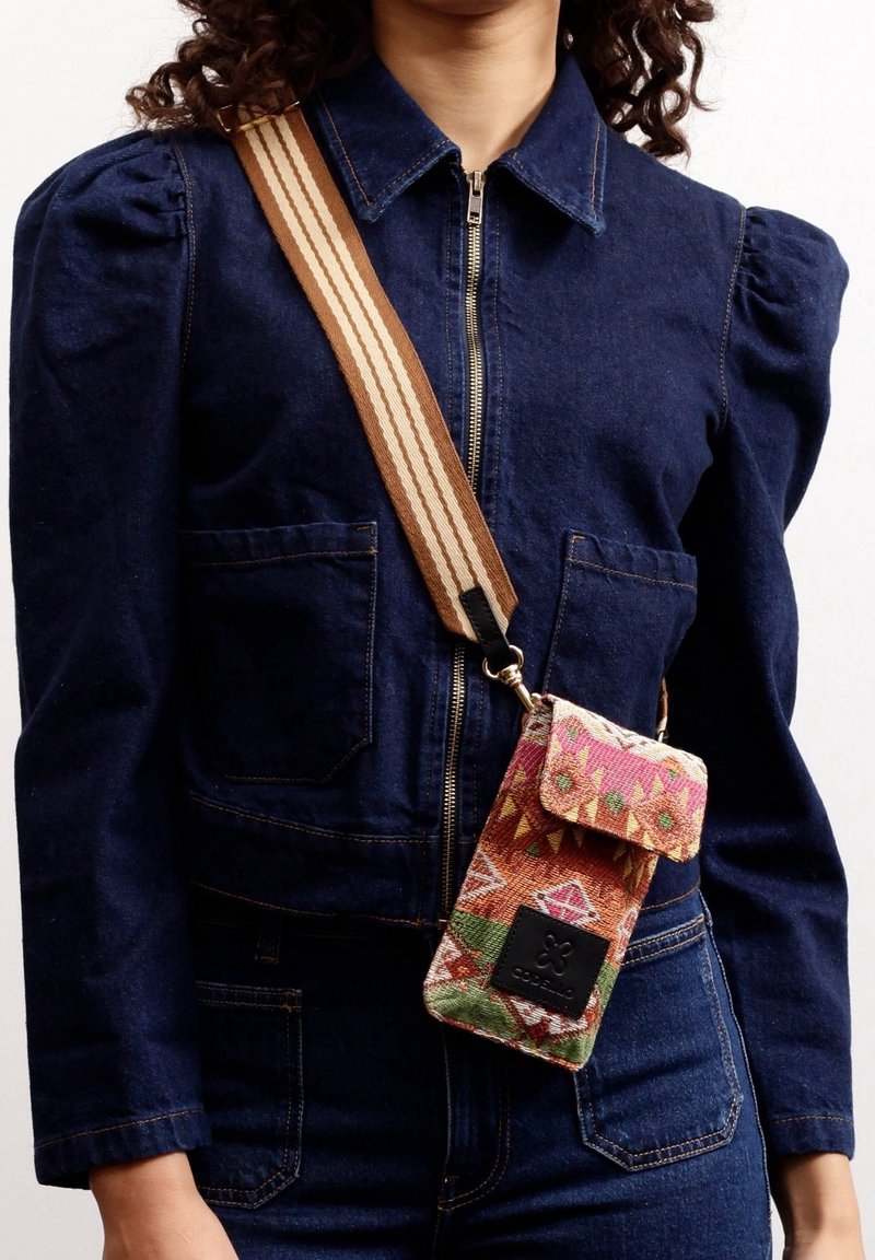 Personne portant une veste en denim bleu foncé et un pantalon avec une pochette pour téléphone à bandoulière colorée et à motifs, avec une large sangle rayée.