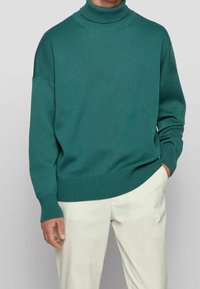 Pull à col roulé côtelé teal porté avec un pantalon blanc cassé, une main dans la poche, visible du cou jusqu'au milieu de la cuisse sur un fond neutre.