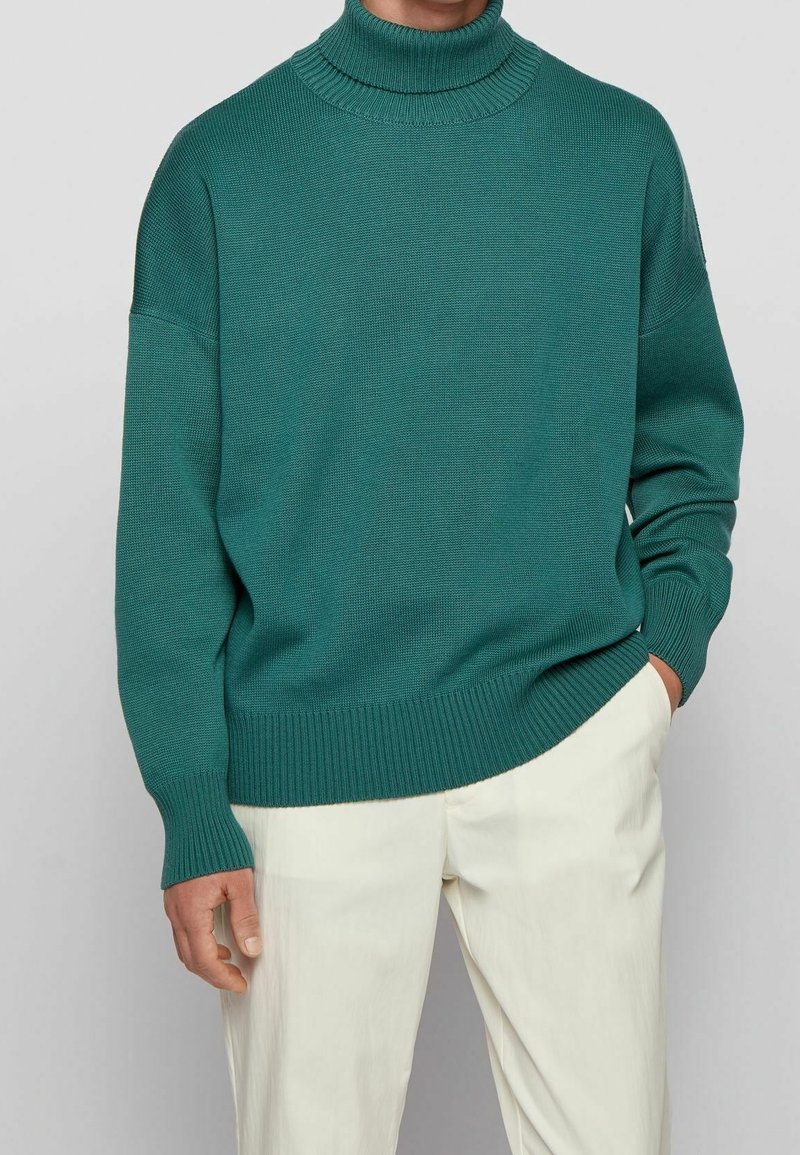 Pull à col roulé côtelé teal porté avec un pantalon blanc cassé, une main dans la poche, visible du cou jusqu'au milieu de la cuisse sur un fond neutre.