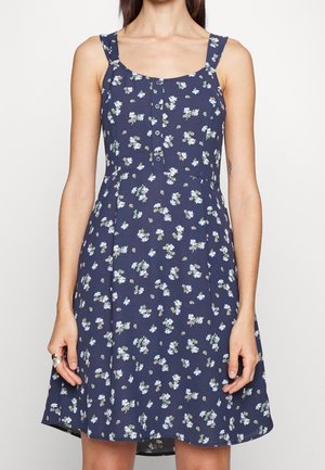 Nainen, joka pukeutunut hihattomaan tummansiniseen mekkoon, jossa on valkoinen ja vaaleanvioletti kukkakuvio sekä edessä napit, seisoo vaaleaa taustaa vasten.