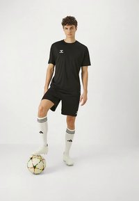 Juoda sportinė marškinėliai ir šortai, baltos dryžuotos per kelį aukštos kojinės, stovint ant futbolo kamuolio, demonstruojant elegantišką, sportišką dizainą.