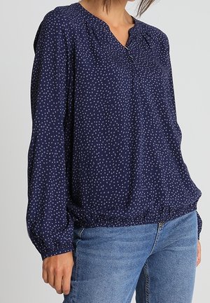 Femme portant un chemisier bleu marine à manches longues avec de petits motifs circulaires blancs et un jean bleu, debout devant un fond uni.