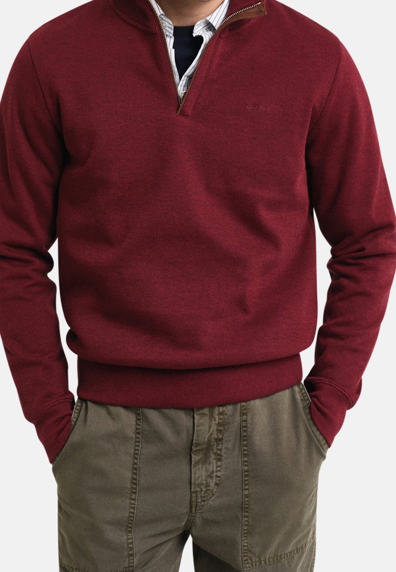 Homme portant un pull à demi-fermeture éclair bordeaux par-dessus une chemise rayée, les mains dans les poches d'un pantalon vert olive, torse visible sur fond uni.