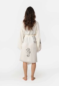 Biele rúcho s čiernym vyšívaním v tvare morského koníka, dlhé rukávy a opasok na zaviazanie. Má textúrovanú látku a franjový lem.